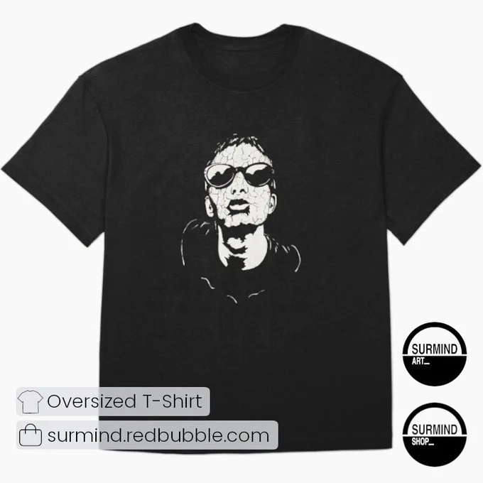 A flat-lay view of a black oversized t-shirt featuring a high-contrast white ink drawing of Thom Yorke in the center. The portrait showcases him looking slightly upward, wearing dark round sunglasses, with his skin textured in a cracked, stylized pattern. To the bottom right, two circular black and white "SURMIND" shop logos are displayed against the light grey background.