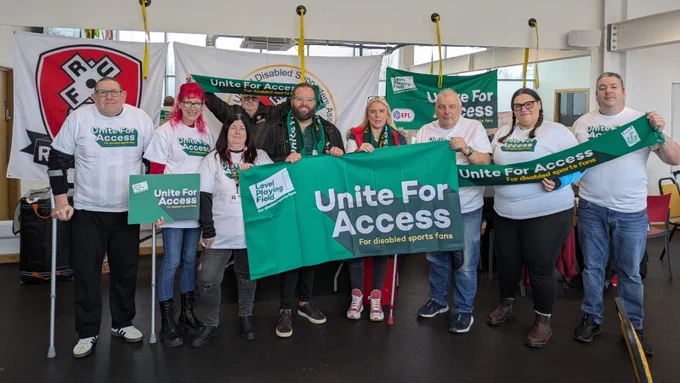 Rotherham United Disabled Supporters' Association members are joined by Level Playing Field's Owain Davies. Everyone is smiling towards camera, with 'Unite For Access' banners, boards, scarves and t-shirts.