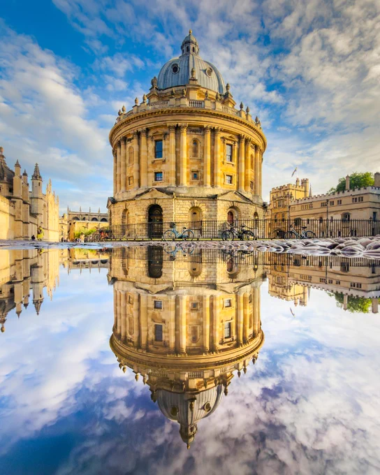 The Radcliffe Camera in central Oxford is reflected almost perfectly in a large puddle after rain. The historic circular building is illuminated by warm sunlight beneath a blue sky scattered with clouds, creating a symmetrical mirror image across the water.