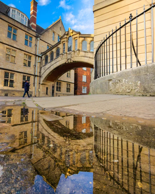 Hertford Bridge, often known as the Bridge of Sighs, spans a narrow street in Oxford. A puddle in the foreground reflects the stone bridge and surrounding college buildings, while a pedestrian walks nearby on a bright day.