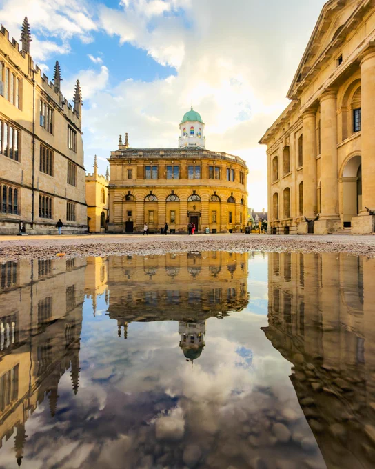 A puddle reflects the Sheldonian Theatre and surrounding historic buildings in central Oxford. Golden sunlight illuminates the stone architecture as a few people cross the open square beneath a partly cloudy sky.