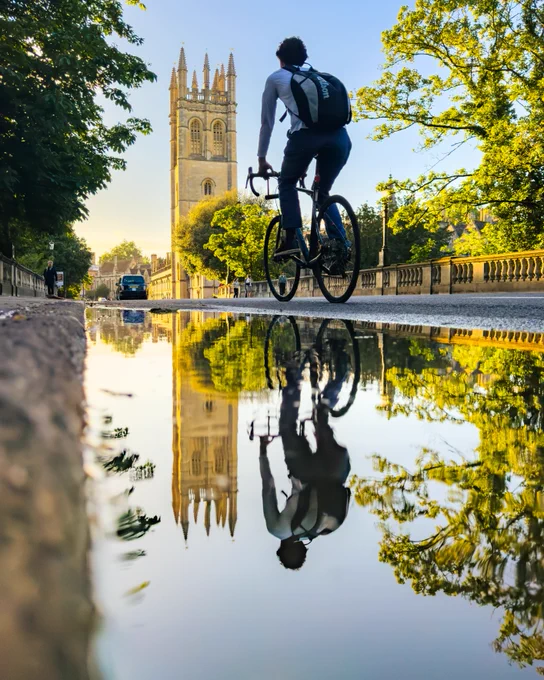 A cyclist rides along a tree-lined Oxford street with Magdalen Tower rising in the background. A large puddle in the road creates a clear reflection of both the cyclist and the Gothic tower in the morning light.