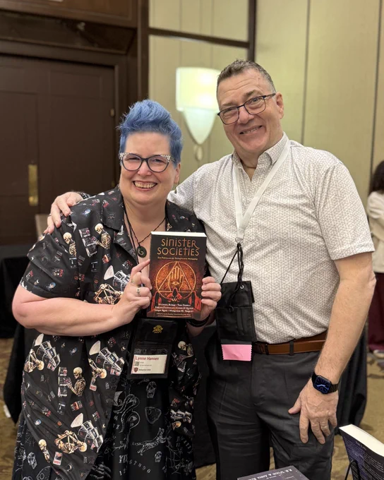 Photo of R. B. Wood with Lynne Hansen holding a paperback copy of SINISTER SOCIETIES, edited by Linda Hartley.