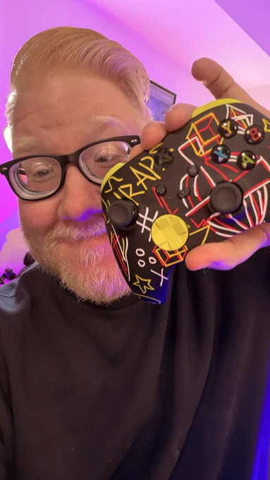 Selfie of me holding up an Xbox controller that has a bunch of Quiggle lines done in a graffiti style in multiple different colours from green to red to white to black with green button, shoulder buttons and D pad and black thumb sticks and other buttons