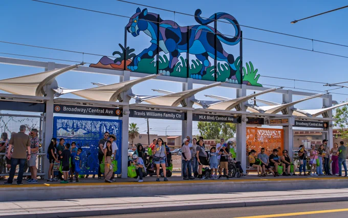 Riders waiting at a light rail station for the light rail to arrive. Above the station is colorful artwork statue depicting an animal and grass. 