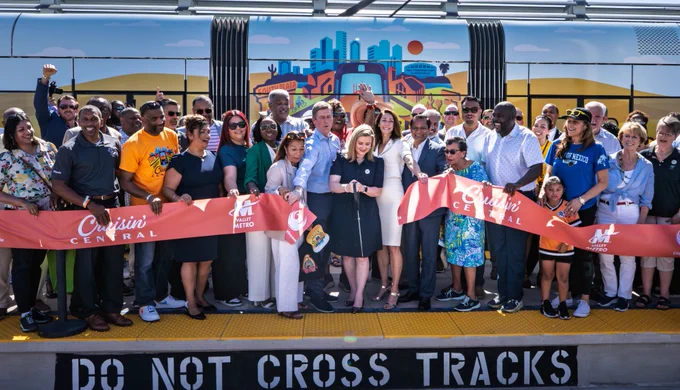 Mayor Kate Gallego cutting a large red ribbon with large scissors, surrounded by other city leadership and community members. They are standing on a light rail platform with a light rail train behind them. 