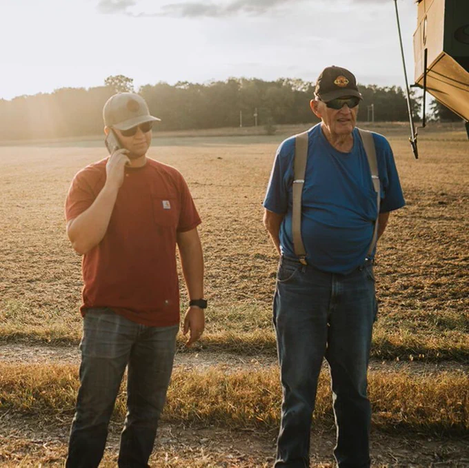 Two generations of farmers stand next to a field while one talks on the phone. 