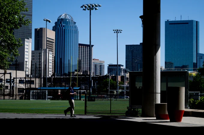 A person walks on a paved path next to a green sports field with goalposts. Several tall, modern city buildings, including one with a domed top and another of reflective blue glass, stand in the background under a clear sky. Bright sunlight casts strong shadows from foreground structures.