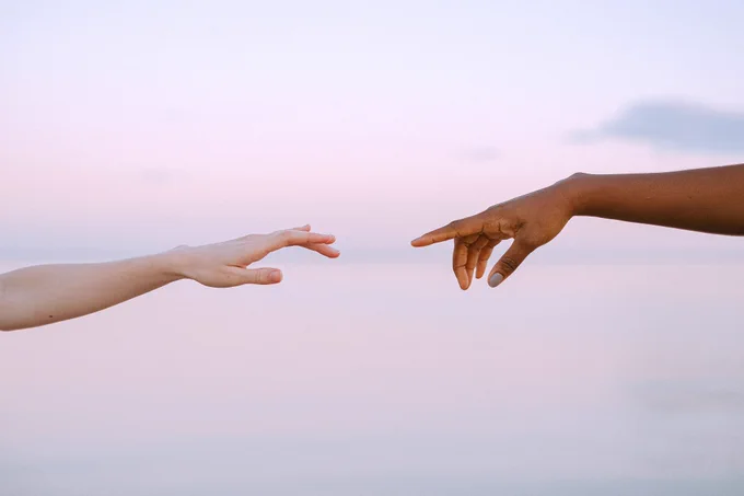 We see a beautiful and peaceful photograph of two hands reaching out towards each other against a landscape of soft pink and purple tones. 