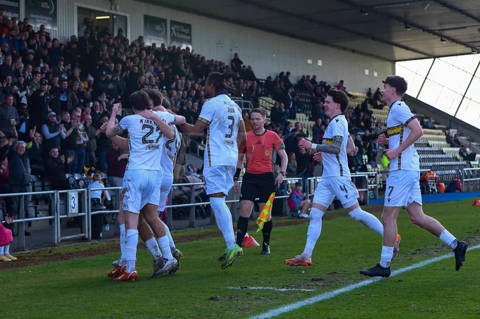 A soccer team in white jerseys jubilantly celebrates a goal during a match. The stands are filled with cheering fans, creating an energetic atmosphere.