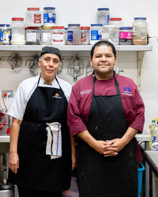 Dos personas vestidas con uniformes de cocina, de pie en una cocina comercial. La persona a la izquierda lleva una chaqueta blanca y delantal negro, mientras que la persona a la derecha viste una chaqueta roja con el texto "Chef Pastelero" y un delantal negro. Están frente a una estantería con recipientes de ingredientes.
