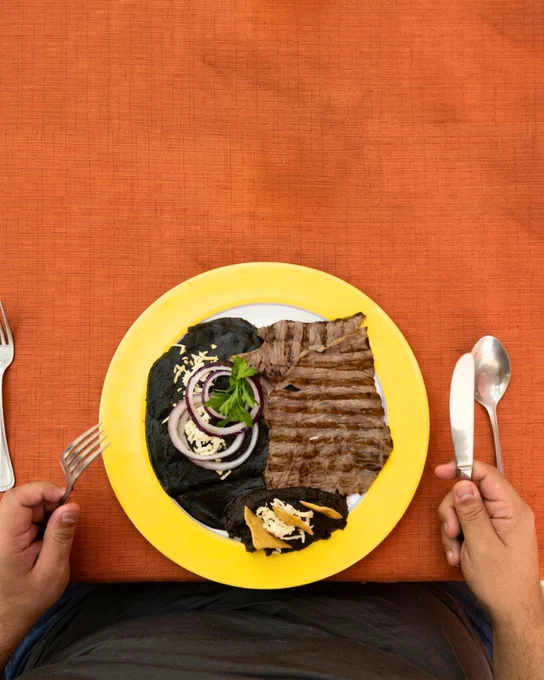 Plato colorido sobre mantel naranja, con enchiladas negras cubiertas de queso y cebolla morada, acompañadas de una porción de carne asada. Una mano sostiene tenedor y otra cuchara.