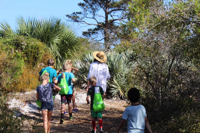 Group of children and an adult walking on a sunny nature trail surrounded by trees and shrubs.