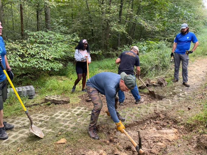 Group of five people working together to dig and clear a dirt path in a forested area using shovels and pickaxes.