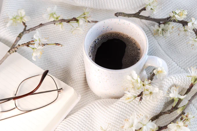 A cup of coffee placed on fabric with tree blossoms and a notebook with glasses on top placed around it