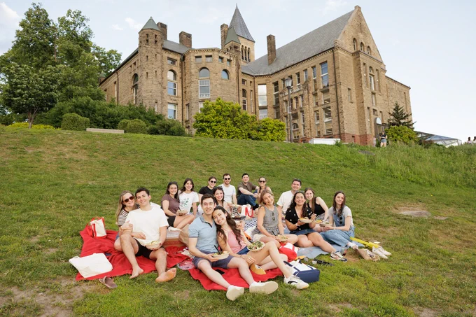 A group of people sitting on Cornell's Libe Slope with Uris Library and McGraw Tower in the background. They are gathered on red blankets, enjoying a picnic. The scene is outdoors with trees and a clear sky in the background.