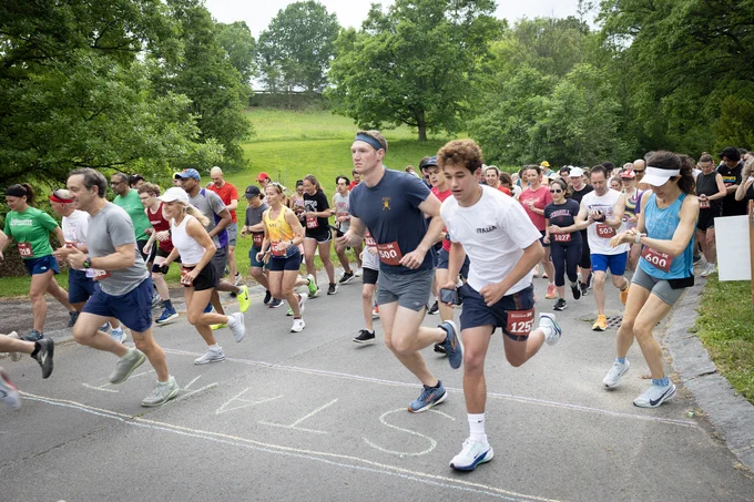 A group of people are running in a park event. They are on a paved path surrounded by green trees and grass. Some runners wear numbered bibs. The path has "START" written in large letters.