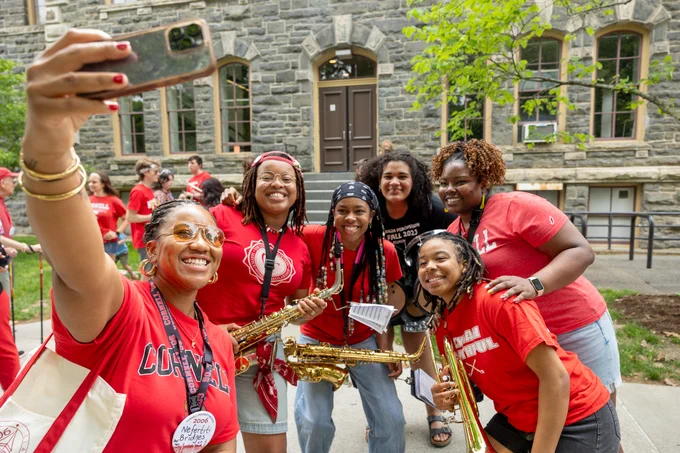 A group of people in red Cornell University shirts pose for a selfie outdoors, with a stone building in the background. Some are holding saxophones, suggesting they are part of a band or musical ensemble. The mood is festive and joyful.