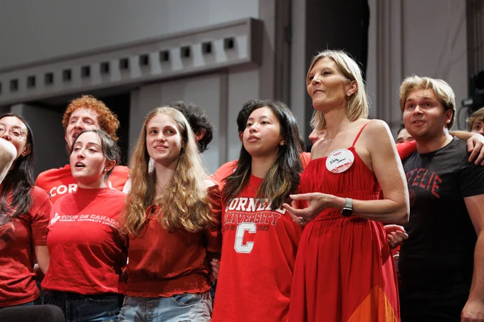A group of people wearing red clothing stand together closely, some with their arms around each other. Several shirts have "Cornell" printed on them, indicating a connection to Cornell University. The background features theater-like decor.