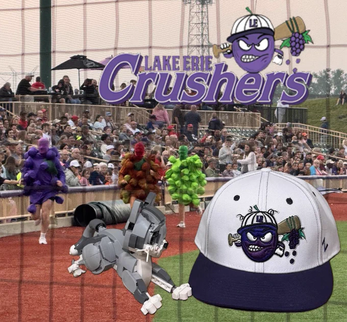 A white and purple Lake Erie Crushers baseball cap with CAPS the Lego Kitten.  A photograph of the grape race is in the background and a Lake Erie Crushers logo is at the top of the image.