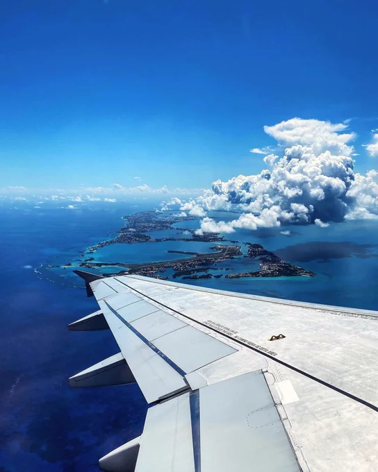Breathtaking airplane window view flying over the vibrant coral reefs and turquoise waters surrounding the island of Bermuda. The airplane wing frames a stunning aerial perspective of the island’s narrow landmass and the L.F. Wade International Airport runway, capturing the magic of arriving at this top-rated island destination.