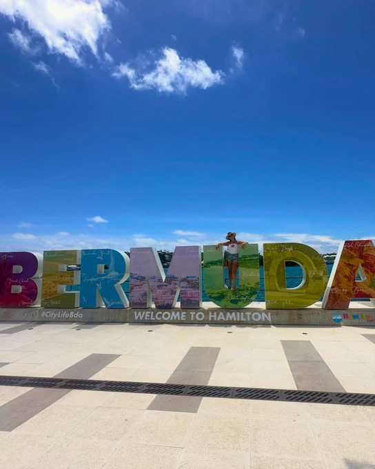 A traveller posing inside the large, colourful "BERMUDA" letter installation on the waterfront terrace, which reads "Welcome to Hamilton." Backdropped by a deep blue harbour and clear sunny skies, this vibrant UGC shot highlights the capital city and invites fans to vote for Bermuda in the Condé Nast Traveller 2026 Readers' Choice Awards.