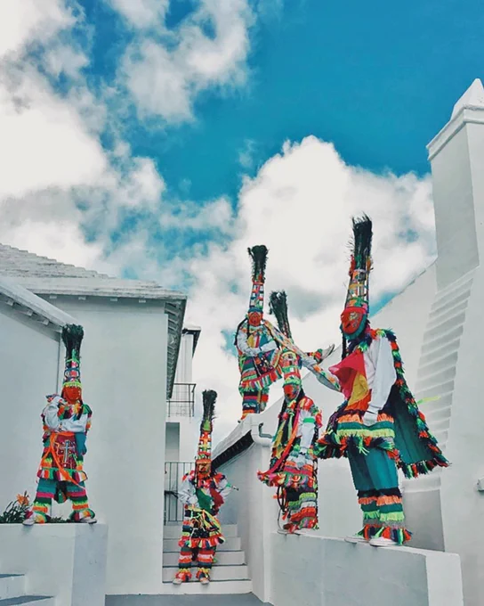 Five traditional Bermudian Gombey dancers dressed in elaborate, colourful fringed costumes, painted masks, and tall feathered headdresses. They are positioned along the white stone steps of a classic Bermudian building with a stepped white roof, showcasing the rich cultural heritage that makes Bermuda a favourite island destination.