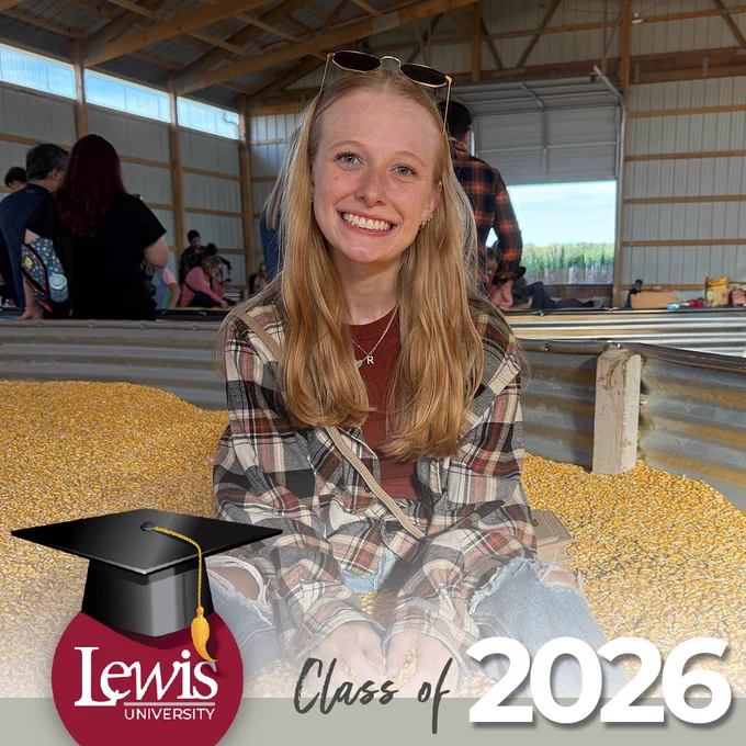 The image shows a smiling person sitting on a pile of yellow corn kernels in a barn or warehouse. The foreground features a graphic with "Lewis University" and "Class of 2026."