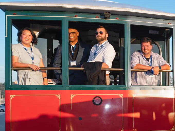 A group of four people smiling and standing on a red Muskegon Trolley car, dressed in business casual attire, wearing name badges. The sun is shining brightly, casting warm light on them.