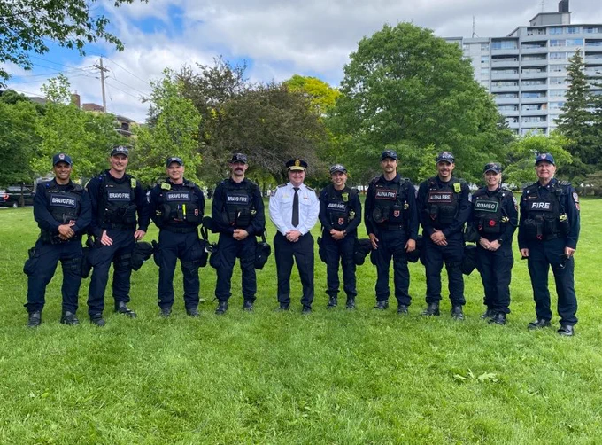 Toronto Fire Chief standing alongside 9 members of the Public Order Unit. Green grass in the forefront and trees and an apartment building in the background.