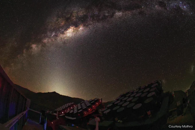 Night sky view featuring the Milky Way galaxy, with an array of telescopes in the foreground. The scene captures a vast expanse of stars and cosmic dust across the sky.