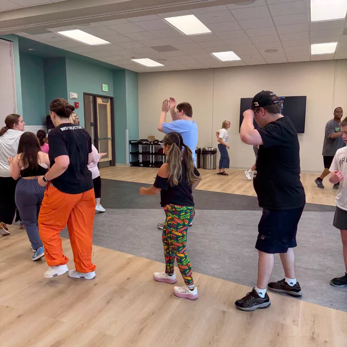 A diverse group of people are participating in a dance or exercise class in a well-lit room. The individuals are standing in a line and appear to be following a routine, with some raising their arms. The room has a wooden floor and pastel-colored walls.