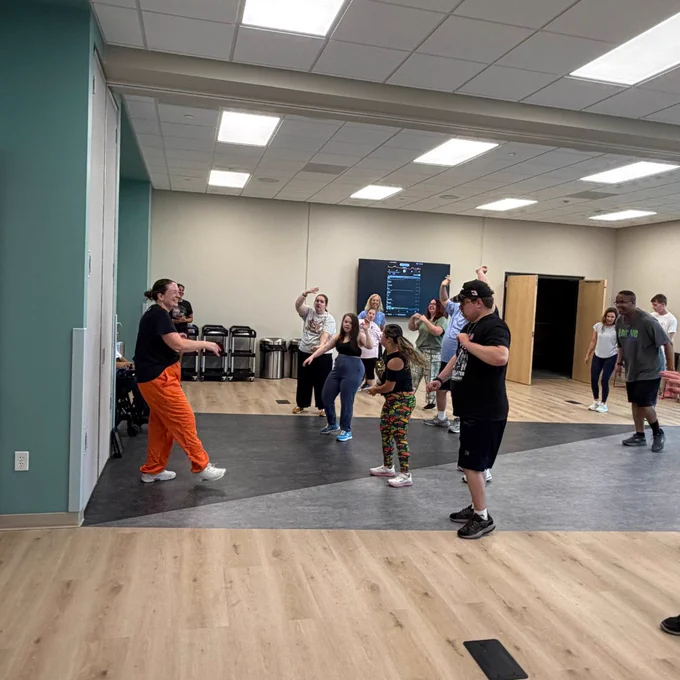 A group of people in a well-lit room, engaging in a lively dance or exercise class. The participants are dressed in casual athletic wear and appear energetic. They are positioned on a wooden floor with a blue accent wall and a screen visible in the background.