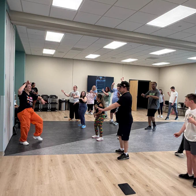 A group of people in a spacious room, actively participating in a dance or exercise class. The instructor at the front is guiding the participants, who are spread out across the room, following along. The environment is well-lit, with a smooth floor and a monitor visible on the wall.