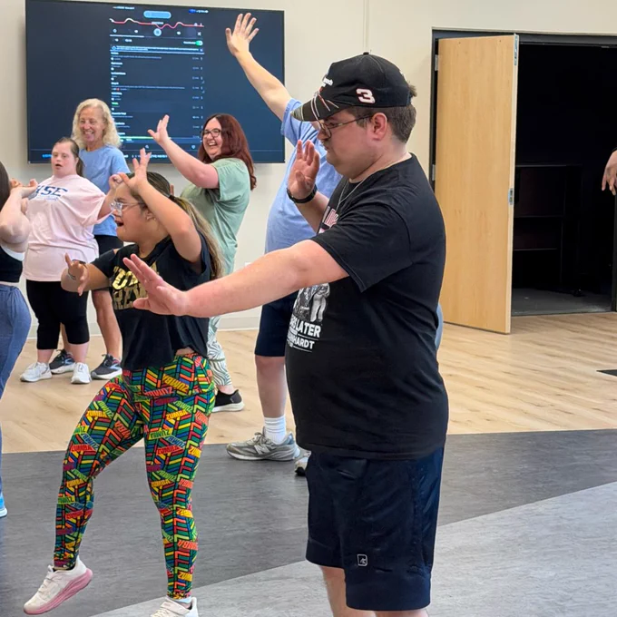 A group of people participating in a choreographed dance class in a room with a wooden floor. A large screen in the background displays information. Participants appear engaged and are following the dance steps, some with their arms raised.