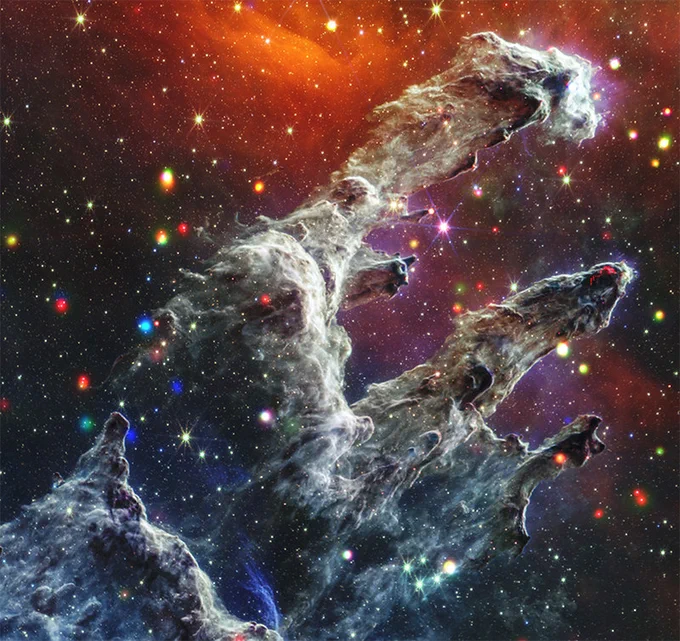 Image showing the "Pillars of Creation" in the Eagle Nebula. Tall, dark, and wispy columnar structures rise against a backdrop of colorful stars and a glowing orange nebula. Bright spots are scattered across the scene.