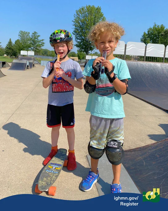 Two boys at a skate park on a sunny day, one wearing a helmet and standing next to a skateboard, the other wearing knee pads and elbow pads.