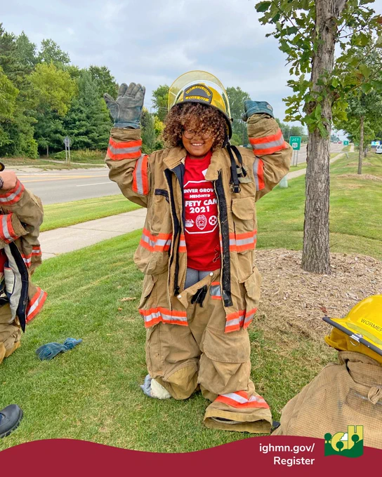 Child dressed in oversized firefighter gear kneeling on grass with hands raised near a roadside tree.