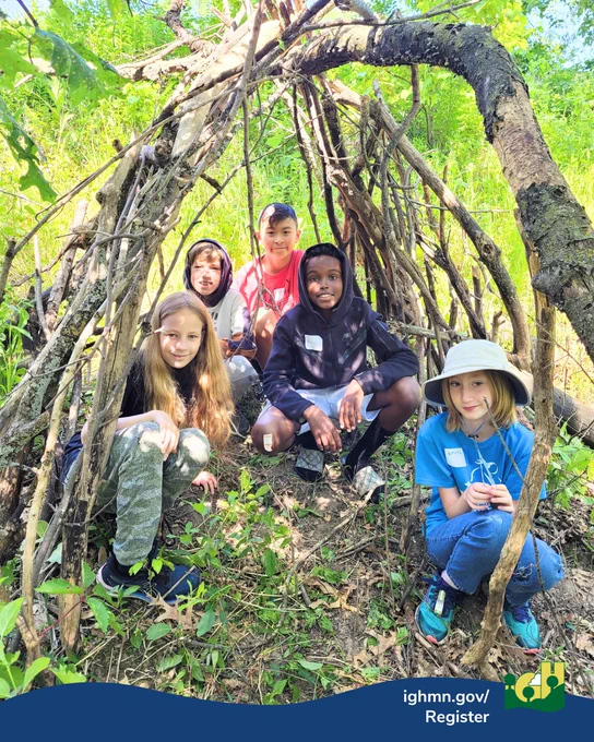 Group of children sitting inside a handmade stick shelter in a sunny forest clearing.