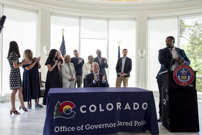 Group of officials applauding behind a table with a Colorado governor's office banner during a formal event.
