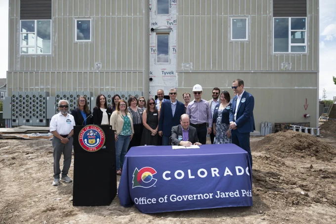 Governor Jared Polis signs a document at a construction site surrounded by officials and staff members in Colorado.