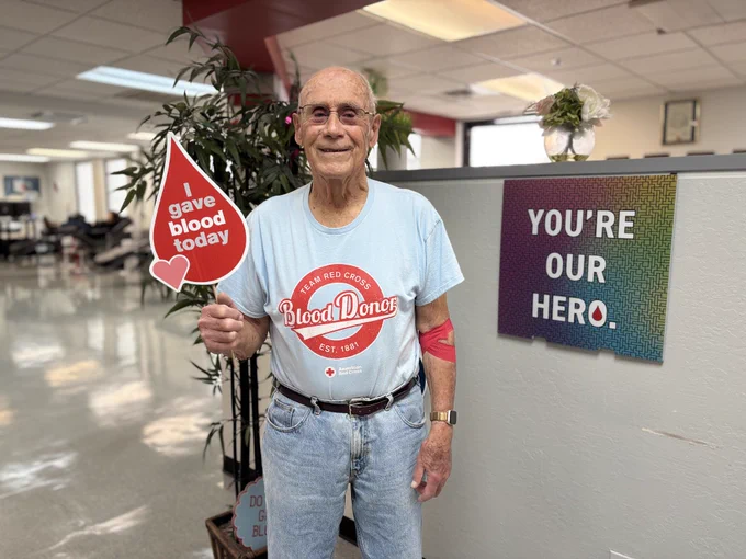 This image shows a person standing in an office environment holding a sign shaped like a blood drop that reads, "I gave blood today." They are wearing a T-shirt with "American Red Cross Blood Donor" on it and have a red bandage on their arm. A sign on a nearby wall says, "You're our hero.