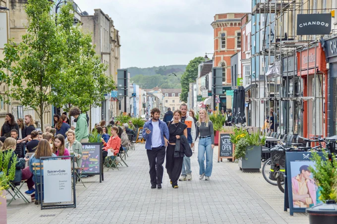 People walk and sit along a pedestrianised shopping street in the centre of Bristol, featuring terraces, trees, planter boxes, bike parking, and advertising boards.