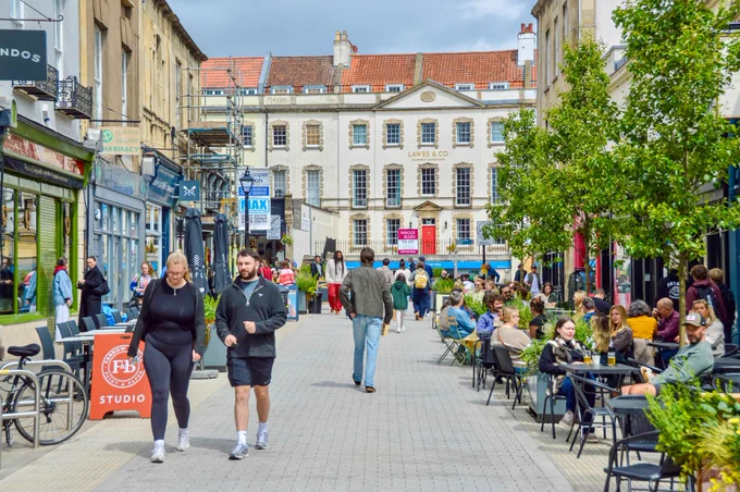 People walk and sit along a pedestrianised shopping street in the centre of Bristol, featuring terraces, trees, planter boxes, bike parking, and advertising boards.