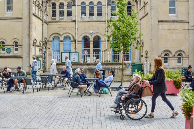 People walk and sit along a pedestrianised shopping street in the centre of Bristol, featuring terraces, trees, planter boxes, bike parking, and advertising boards.