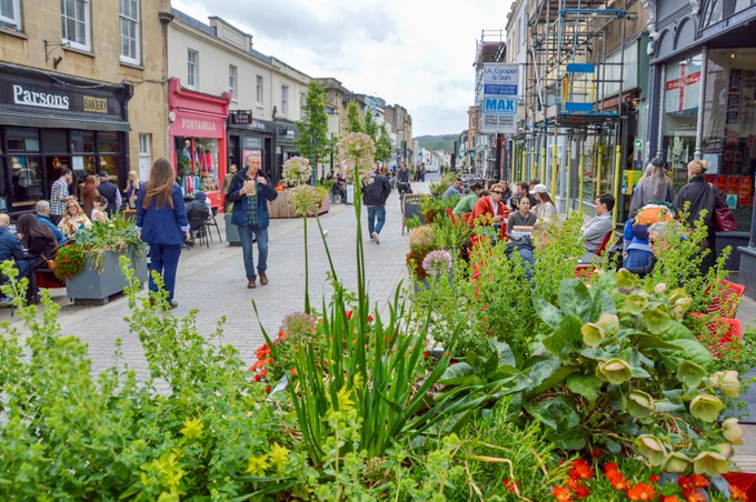 People walk and sit along a pedestrianised shopping street in the centre of Bristol, featuring terraces, trees, planter boxes, bike parking, and advertising boards.