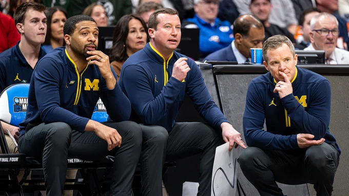 Photo shows assistants Drew Williamson and Kyle Church in Blue Jordan gear with head coach Dusty May on the bench coaching