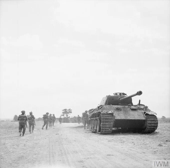 A group of soldiers walk along a dirt road beside a German Panther tank in a rural landscape. The scene is calm, with a few trees visible in the background under a partly cloudy sky.
