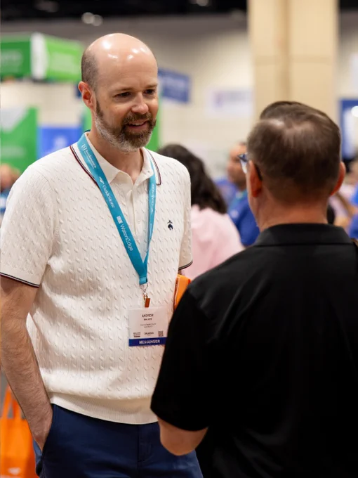 Two men engage in conversation at a professional event. Name tags indicate a formal setting. The atmosphere is one of networking and exchange.