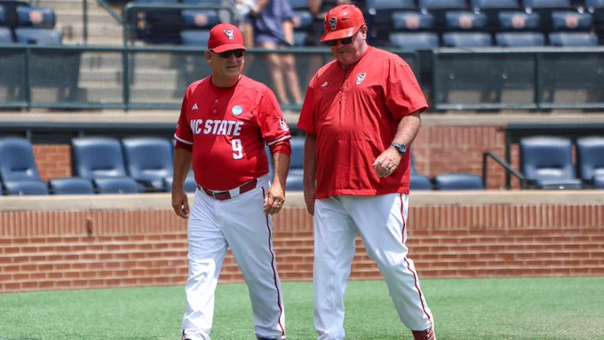 Two NC State baseball coaches in red uniforms walking on the field during a game or practice session.